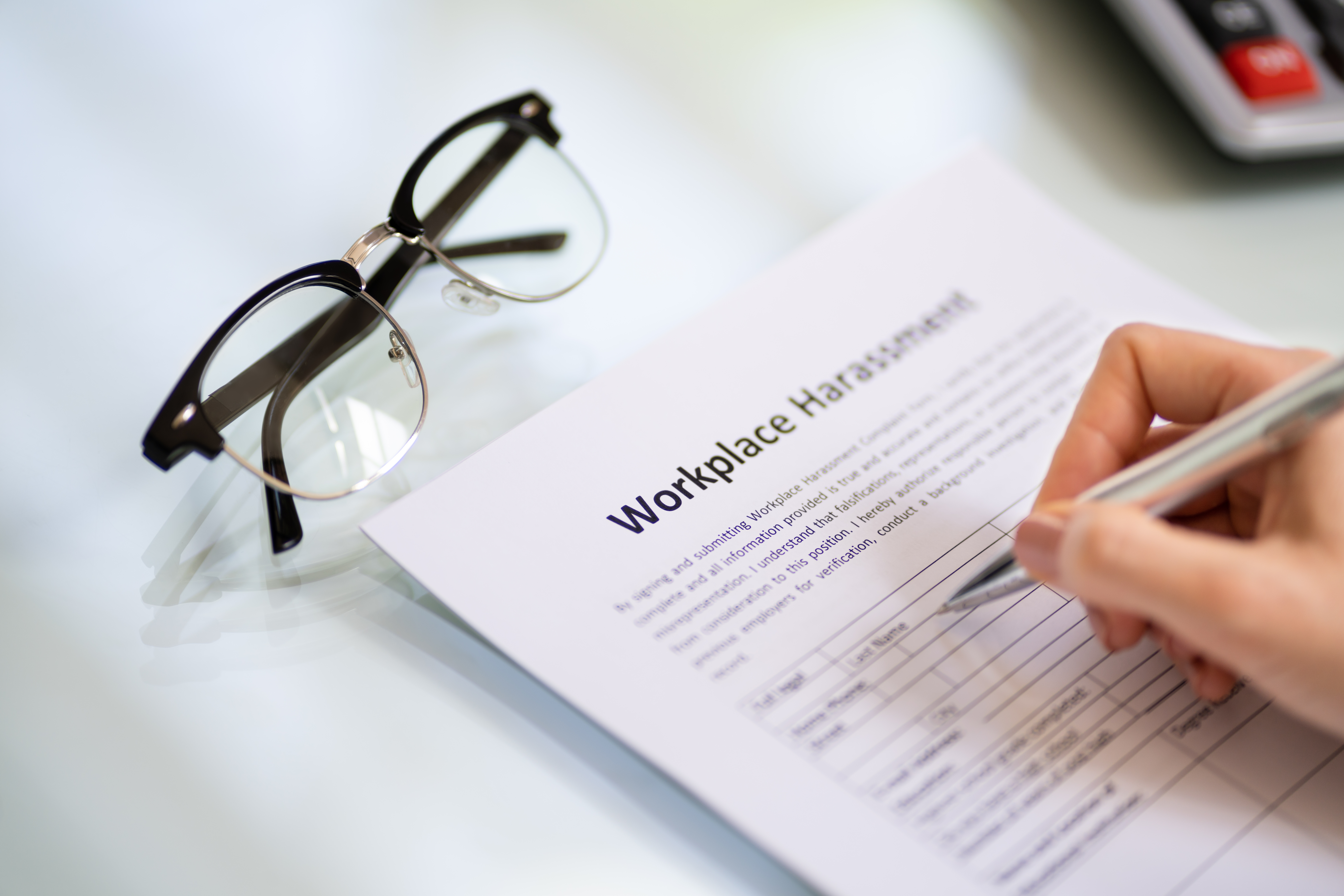 The image depicts a closeup of an employee filling out a workplace harassment document at a desk, listing any forms of harassment that has ben experienced by another individual in the workplace.