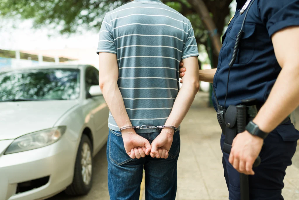 Image depicts a male being arrested by a police officer in front of a car, indicating various reasons for the arrest, such as driving under the influence (DUI) or causing a hit-and-run accident.